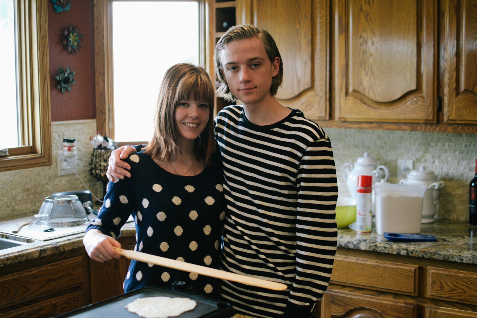 A side image of a woman wearing a navy and white polka dot sweater with her arm around a man wearing a navy and white striped sweater in front of a griddle with potato flatbread.