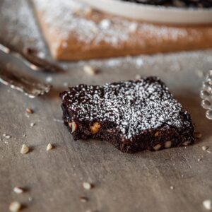 A closeup image of a brownie with a bite taken out of it on a brown counter next to a wood board and chopped hazelnuts.