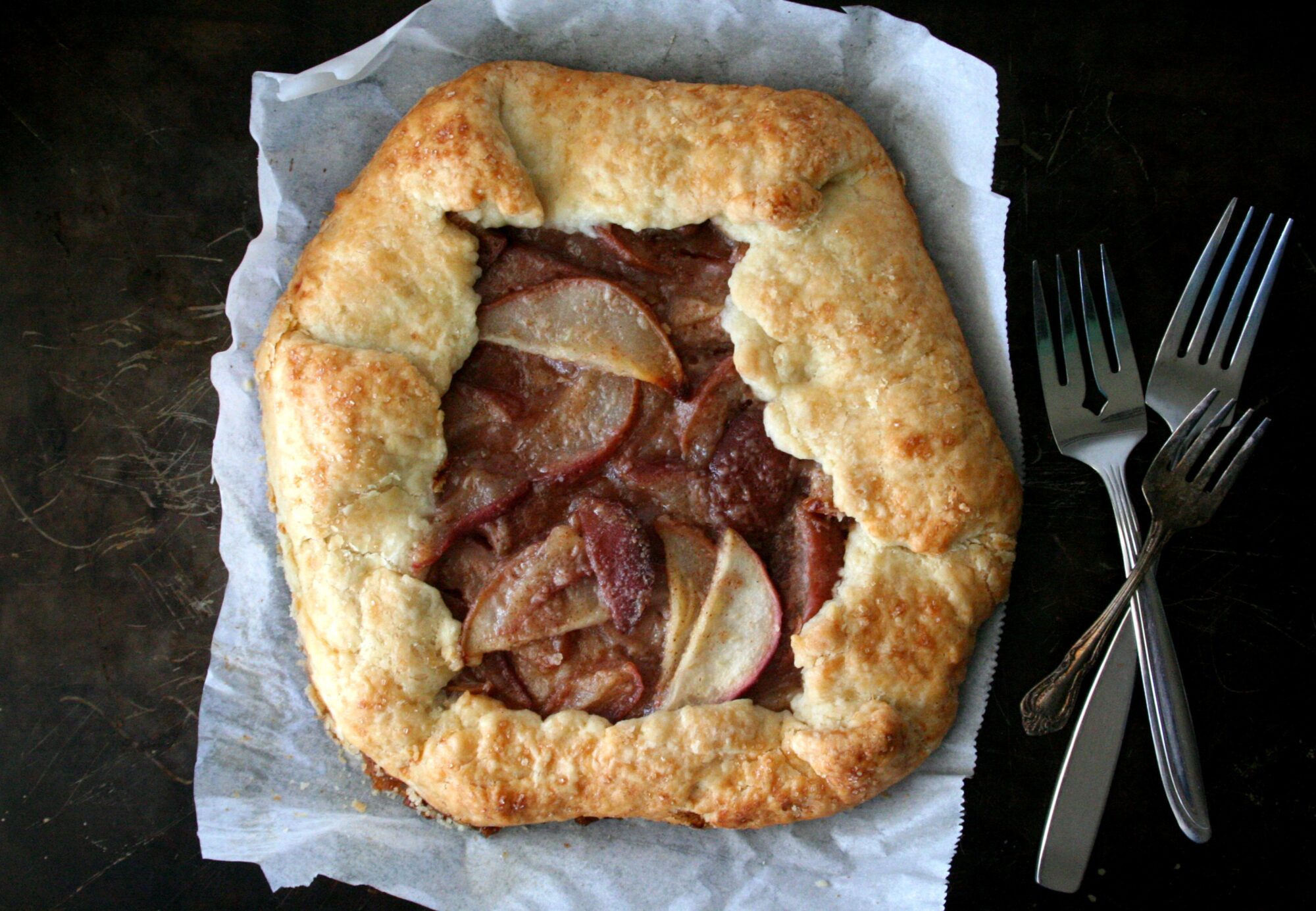 An overhead image of a pear galette on a black sheet pan next to vintage forks.