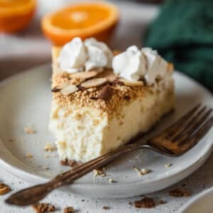 A slice of mascarpone cheesecake with a bite taken out of it on a white plate with oranges and a green linen in the background.