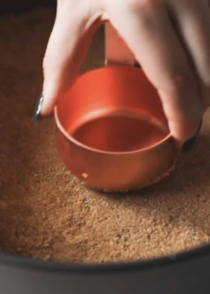 A hand using a measuring cup to press almond cookie crust into a pan.