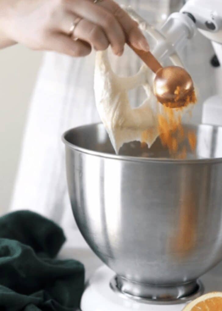 A hand adding orange zest to a silver bowl of filling for mascarpone cheesecake.