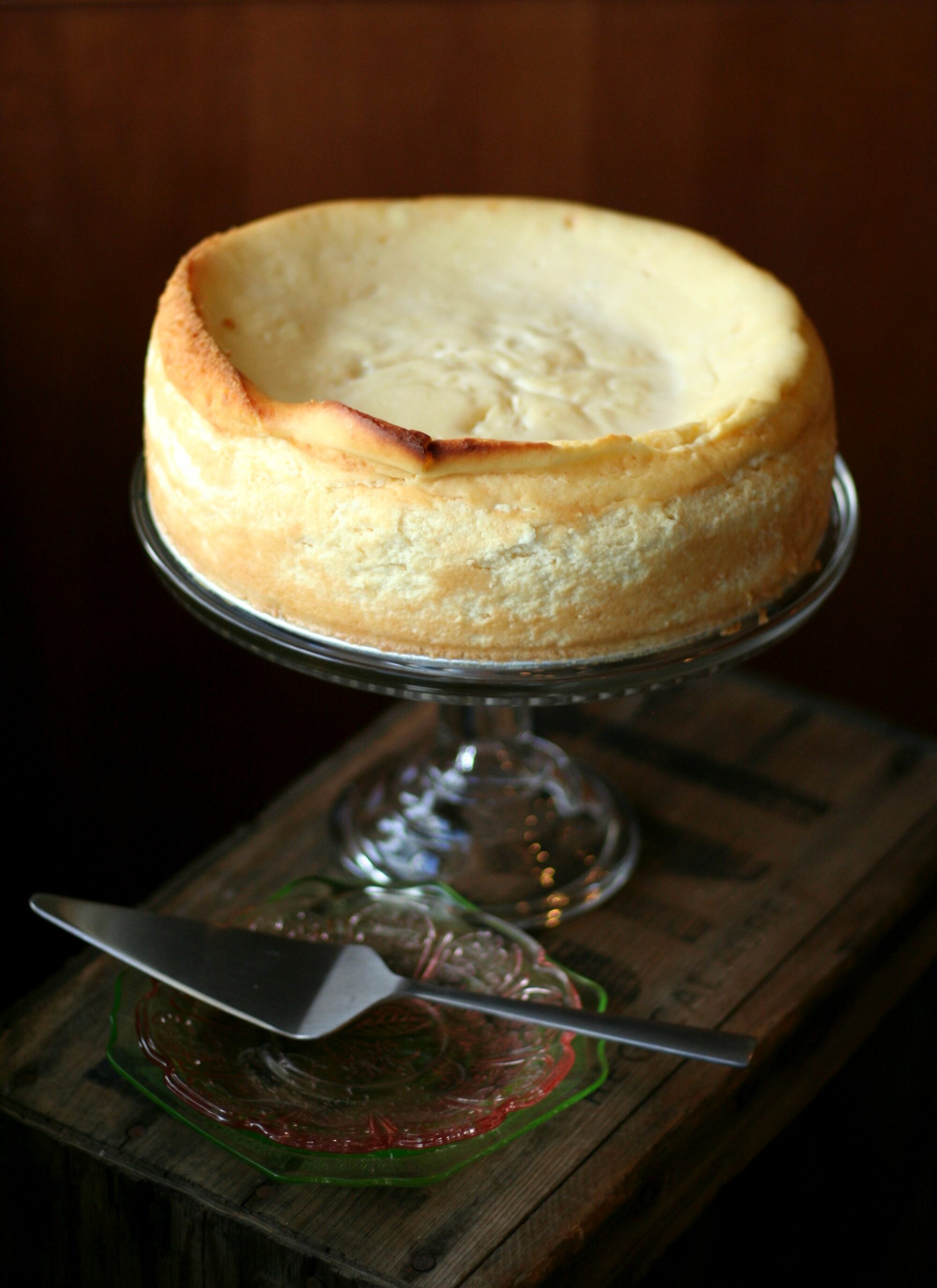 A side image of a cheesecake on a glass stand with a wood background next to vintage green and pink plates.