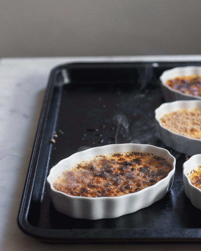 A gif of a white dish of creme brulee with smoke rising off of it in a black sheet pan with a marble background.