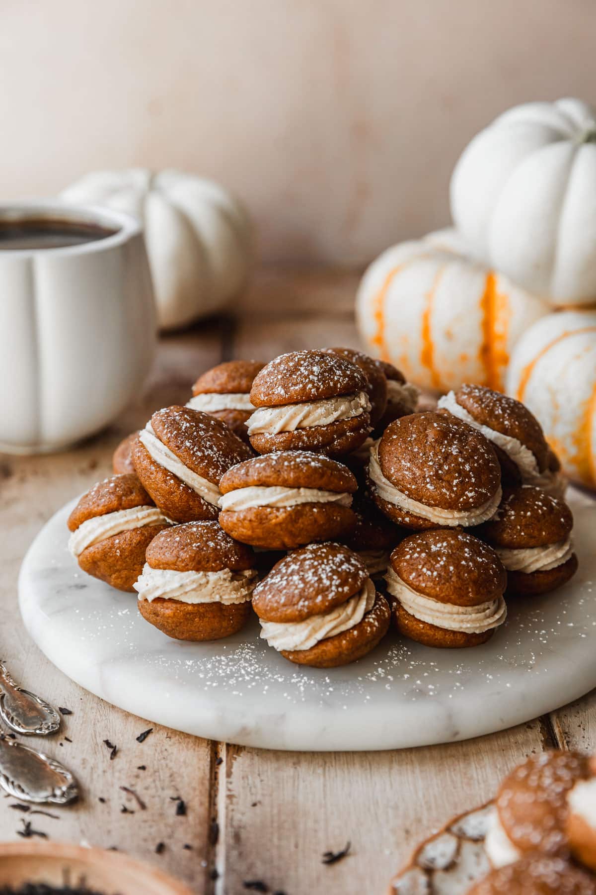 Mini pumpkin whoopie pies with masala chai buttercream on a white tray next to pumpkins, a white mug of coffee, and forks on a wood table.