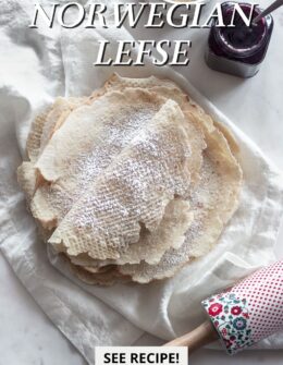 An overhead image of a stack of lefse on a white linen next to a purple jar of jam, white bowl of brown sugar, and red and blue floral rolling pin.