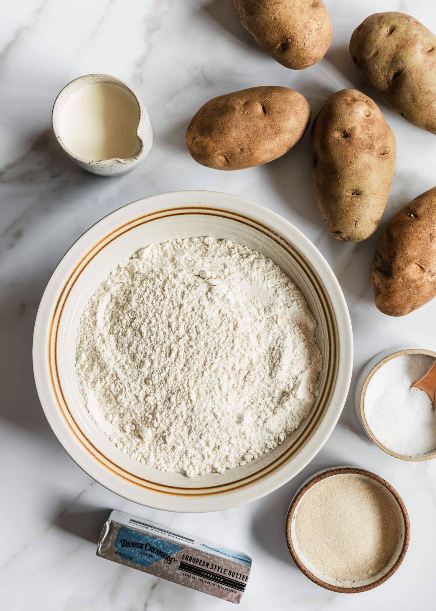 Potatoes, a white bowl of flour, a white bowl of salt, a white bowl of cream, a brown bowl of sugar, and a stick of butter on a white marble counter.