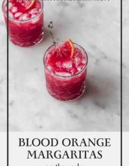 A side image of two blood orange margaritas with orange slices and thyme on a white marble counter.