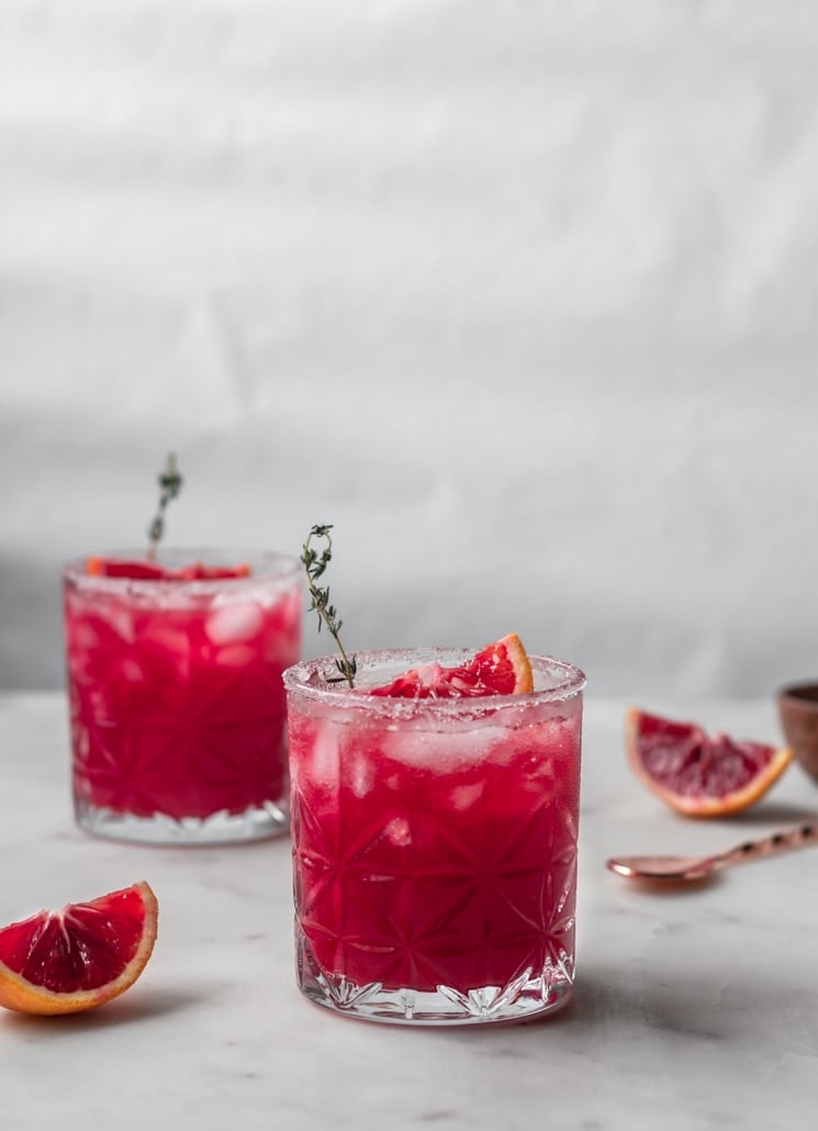 Two pink cocktails garnished with slices of orange and sprigs of thyme on a white counter next to a slice of orange and copper cocktail spoon.