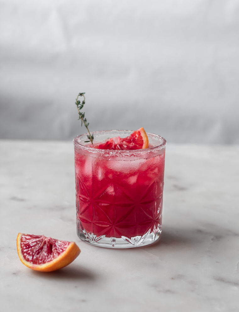 A closeup photo of a blood orange margarita garnished with thyme on a white table next to a slice of orange.