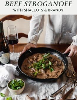 A woman holding a pan of beef stroganoff on a wood table next to a bottle of wine and bowl of parsley.