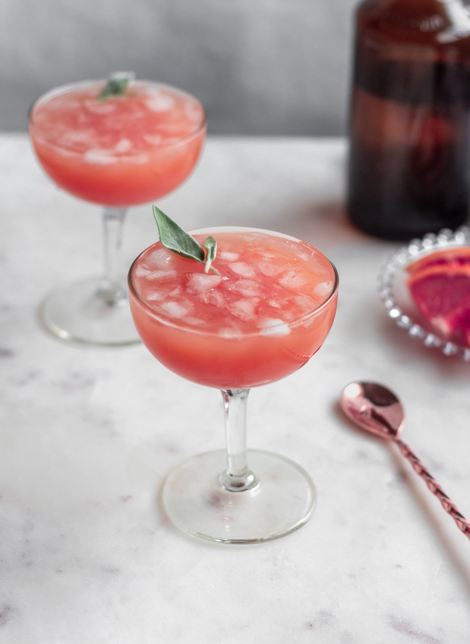 A side image of a sage greyhound cocktail on a marble counter with another cocktail, plate of grapefruit, and copper spoon in the background.