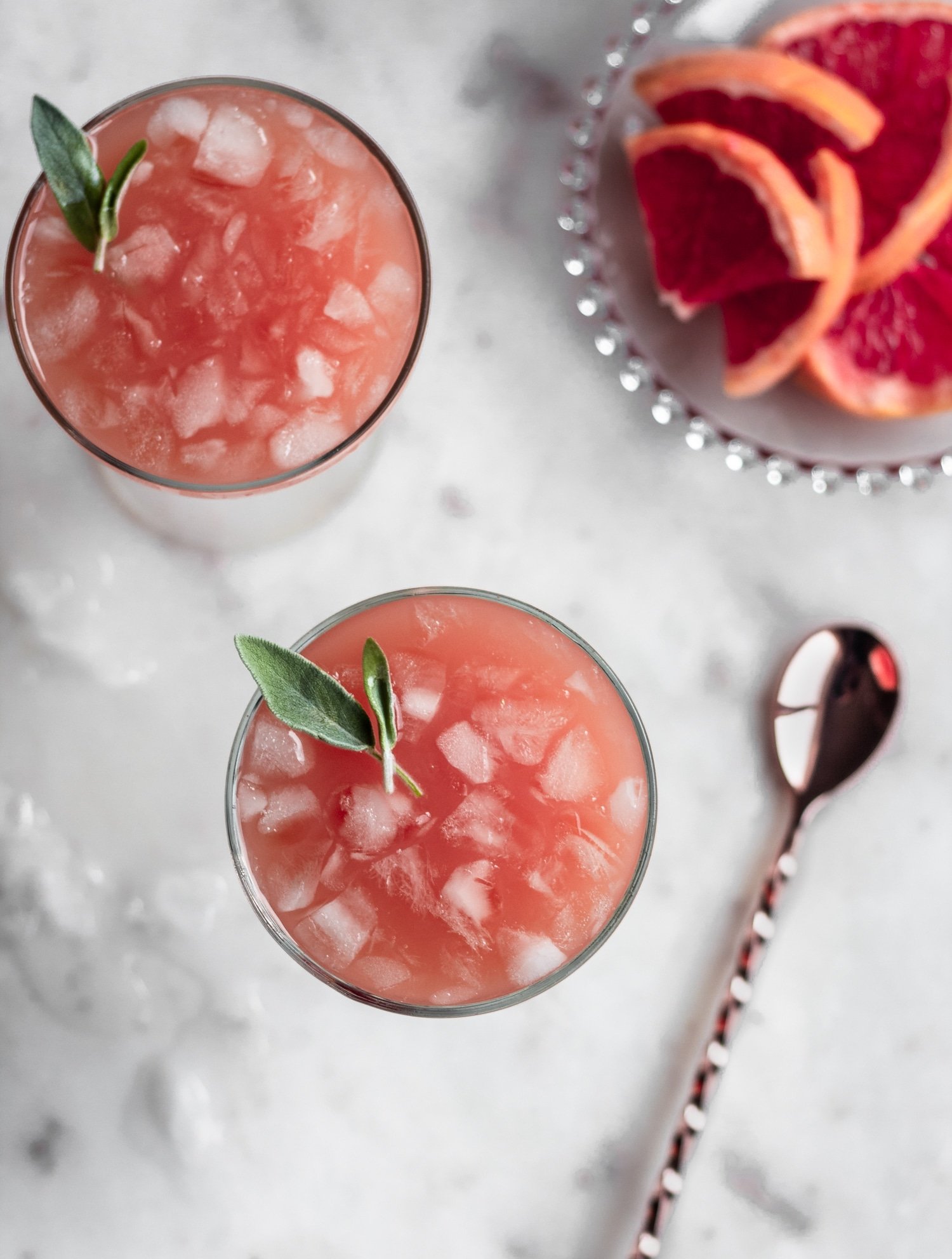 An overhead image of two sage greyhound cocktails garnished with sage leaves on a marble counter next to a plate of grapefruit slices.