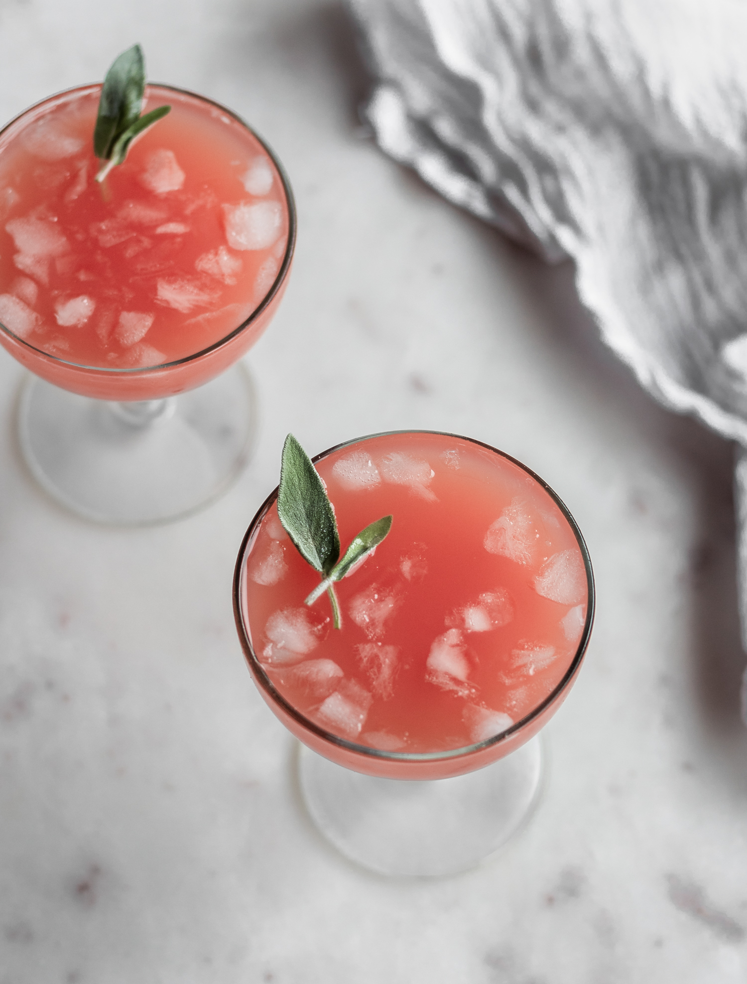 A 45-degree image of two pink cocktails garnished with sage on a marble counter.