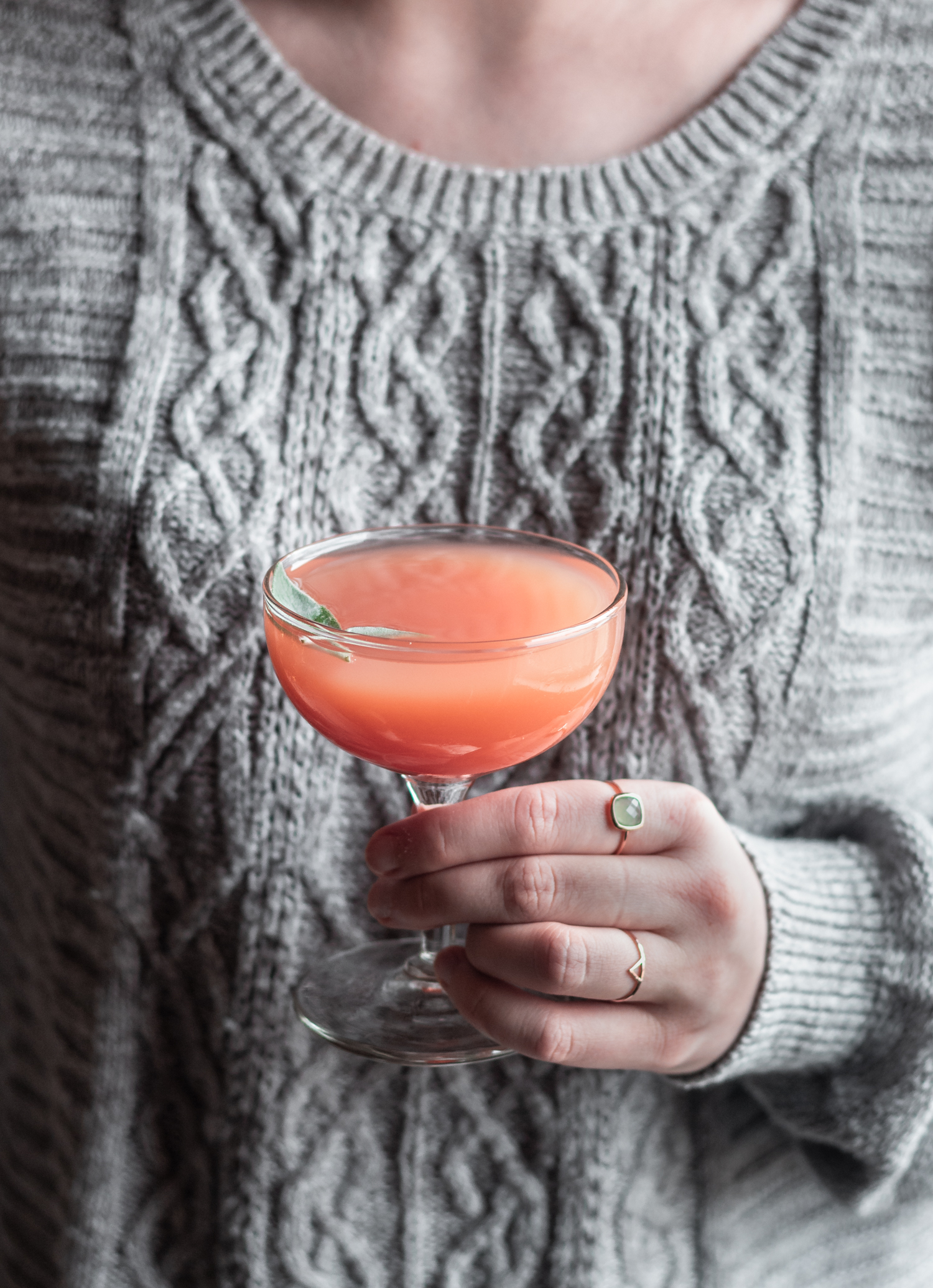 A side image of a woman wearing a grey sweater holding a sage greyhound cocktail in a coupe glass.