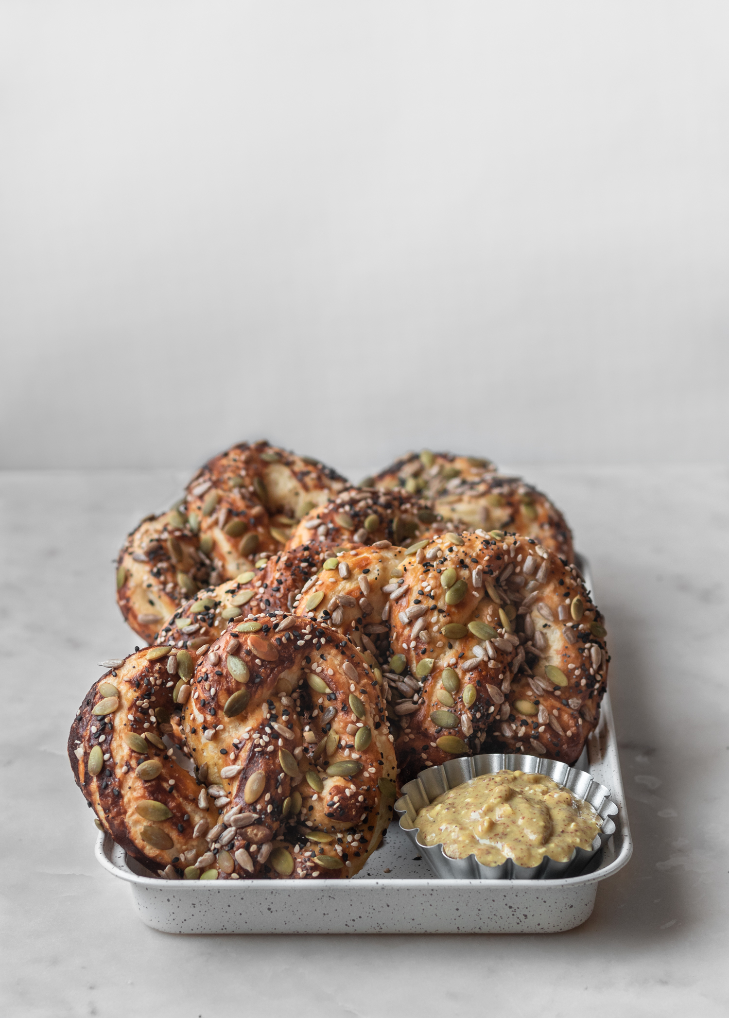 A row of seedy soft pretzels on a white sheet pan, sitting on a marble counter.