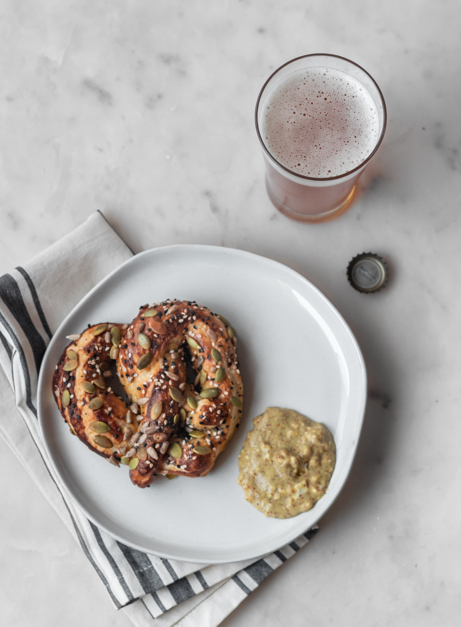 A soft pretzel on a plate sitting on a folded striped napkin on a white background. Glass of beer in the corner.