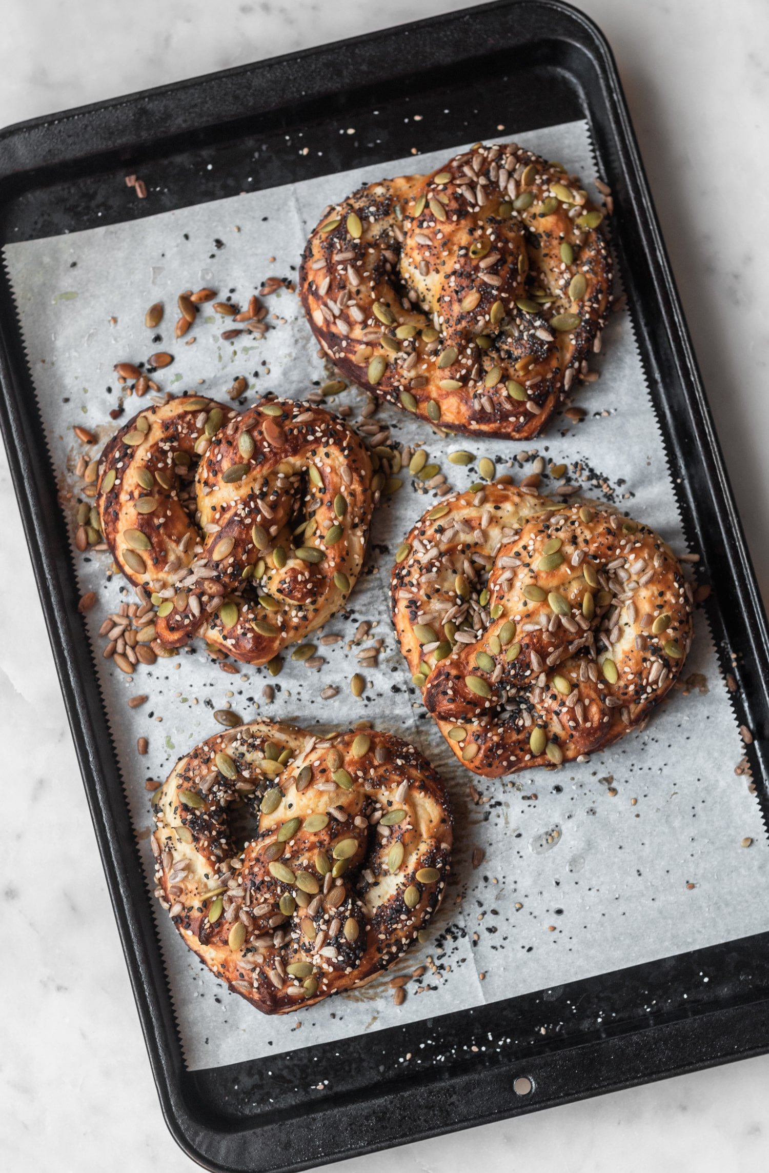 A black sheet pan filled with soft pretzels covered in seeds on a white counter top.