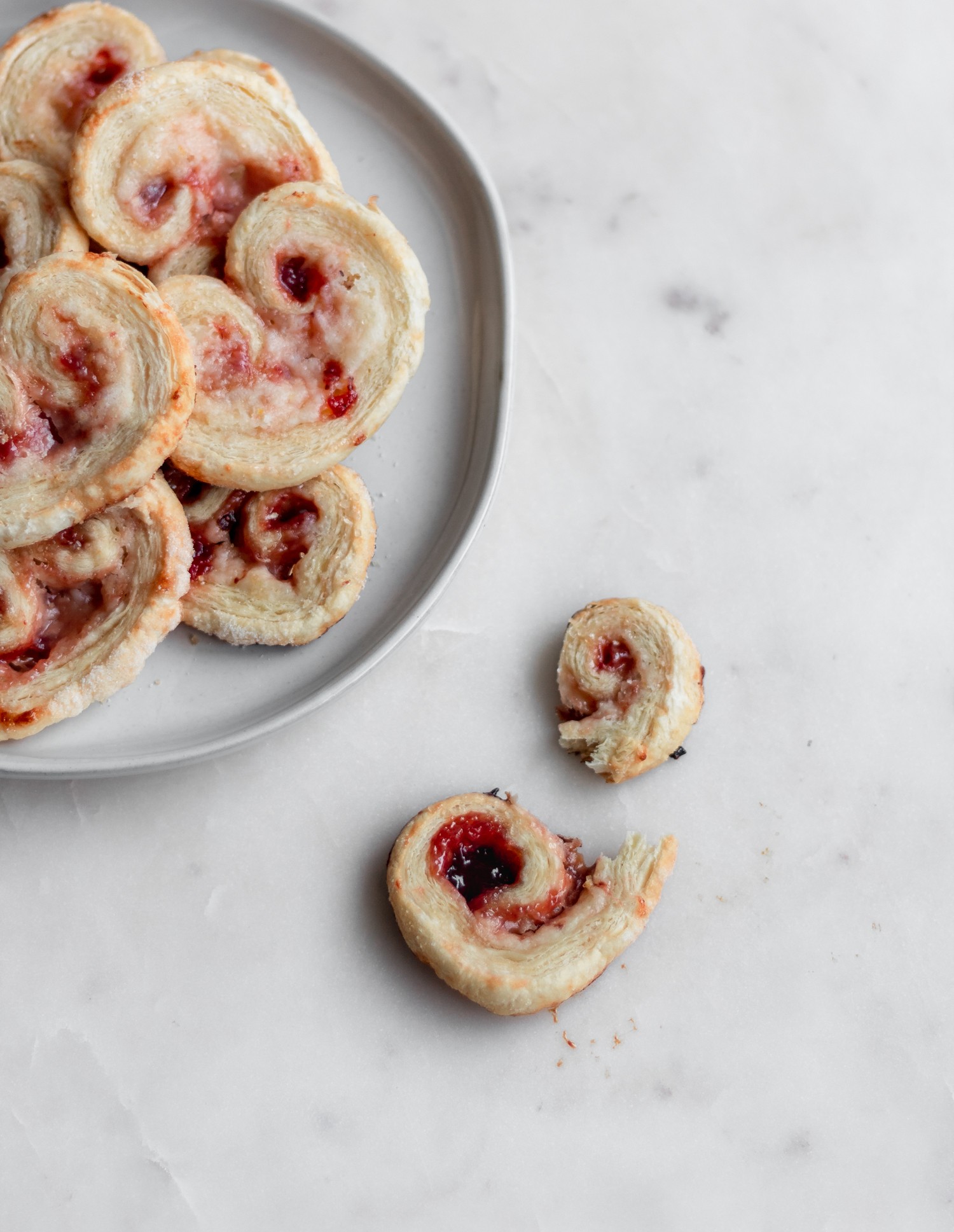 An overhead image of a broken strawberry palmier on a white counter next to a white plate with more cookies.