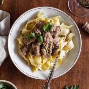 A closeup overhead image of a white bowl of stroganoff with pasta and a parsley garnish on a wood bowl next to a beige linen.