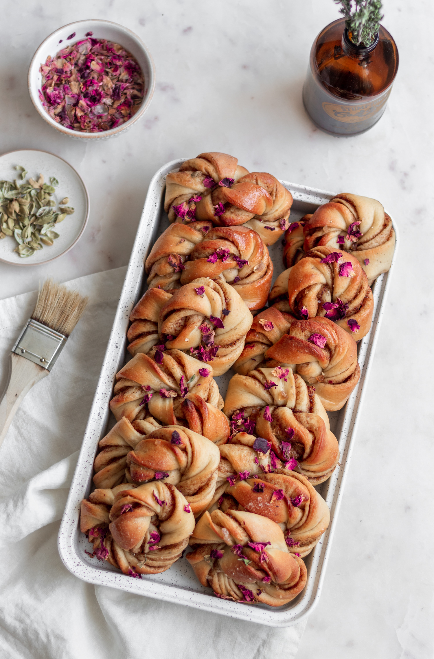 An overhead image of rose cardamom buns in a white tray on a marble counter next to a white linen, white bowl of cardamom pods, and a white bowl of rose petals.