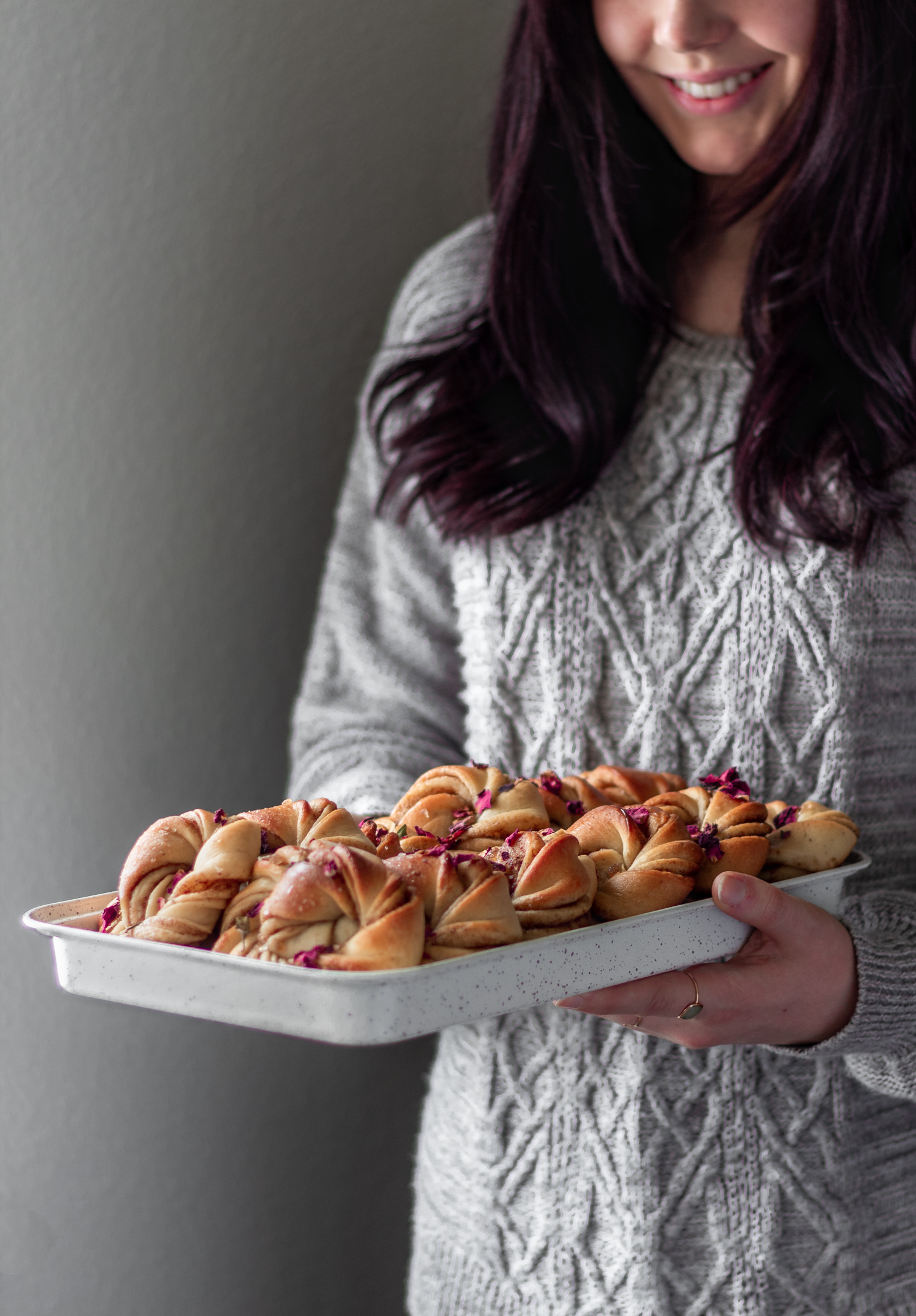 A side image of a woman with dark purple-red hair wearing a grey sweater holding a white tray of Swedish cinnamon buns.