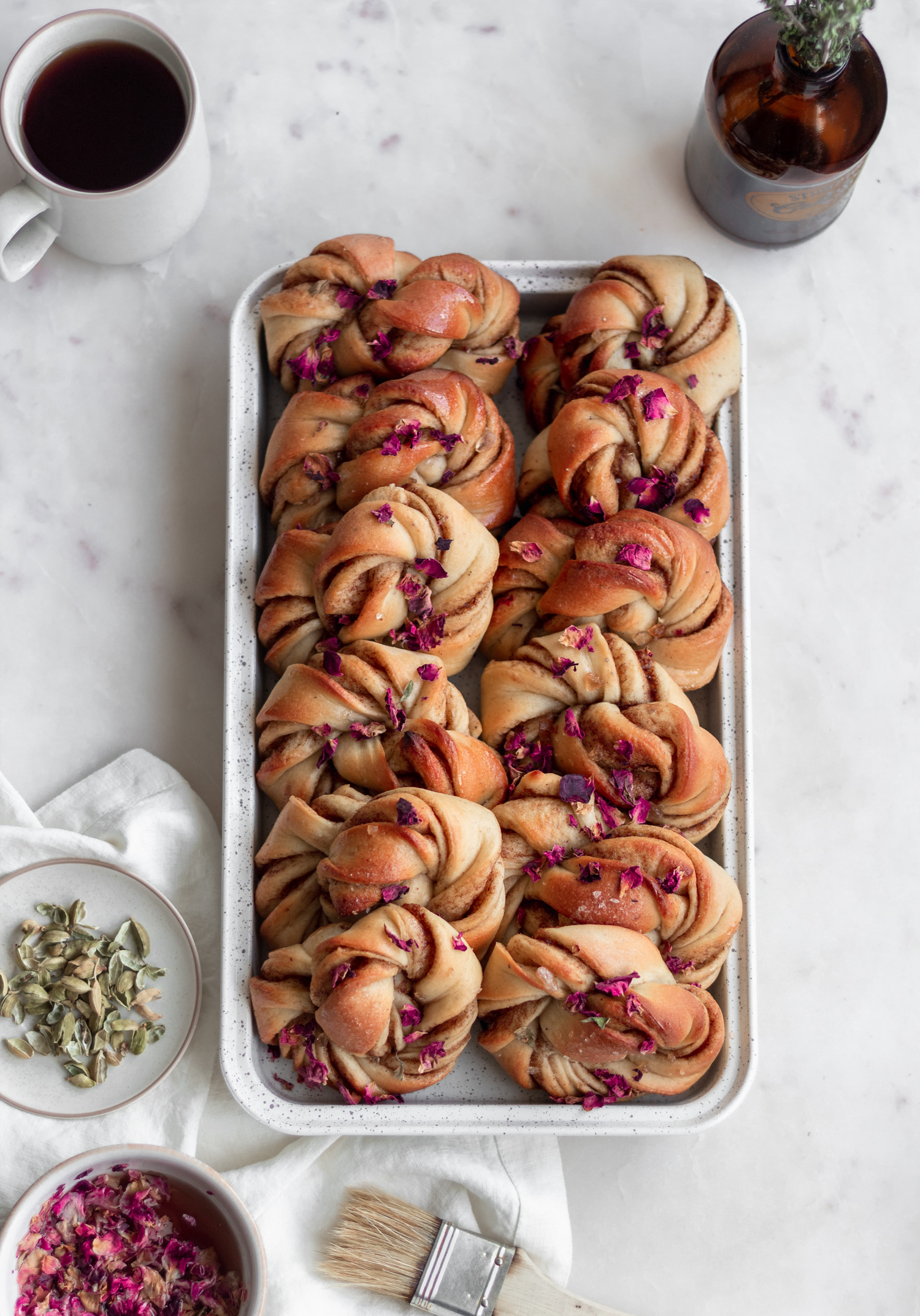 An overhead image of a white baking dish with Swedish cinnamon rolls on a marble counter next to a white linen, cup of coffee, and white plate of cardamom pods.