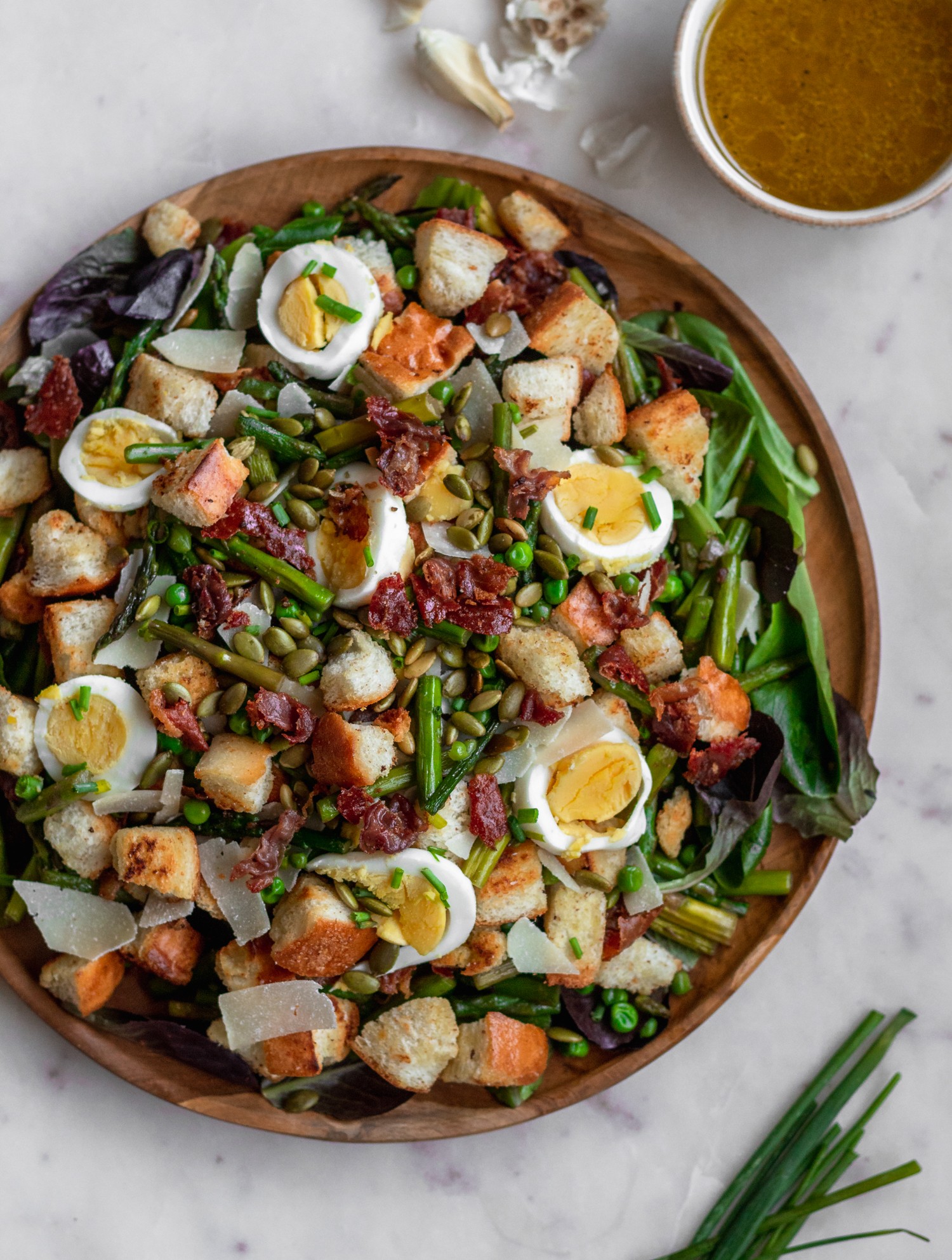 An overhead image of a spring panzanella salad with asparagus, peas, prosciutto, and eggs on a marble counter next to chives and a white bowl of dressing.