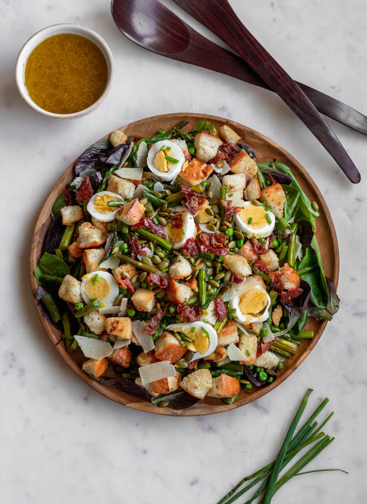 An overhead image of a wood plate with a Spring Panzanella salad with greens, spring veggies, hard boiled eggs, and croutons on a marble counter next to a white bowl of dressing and wood salad tossers.