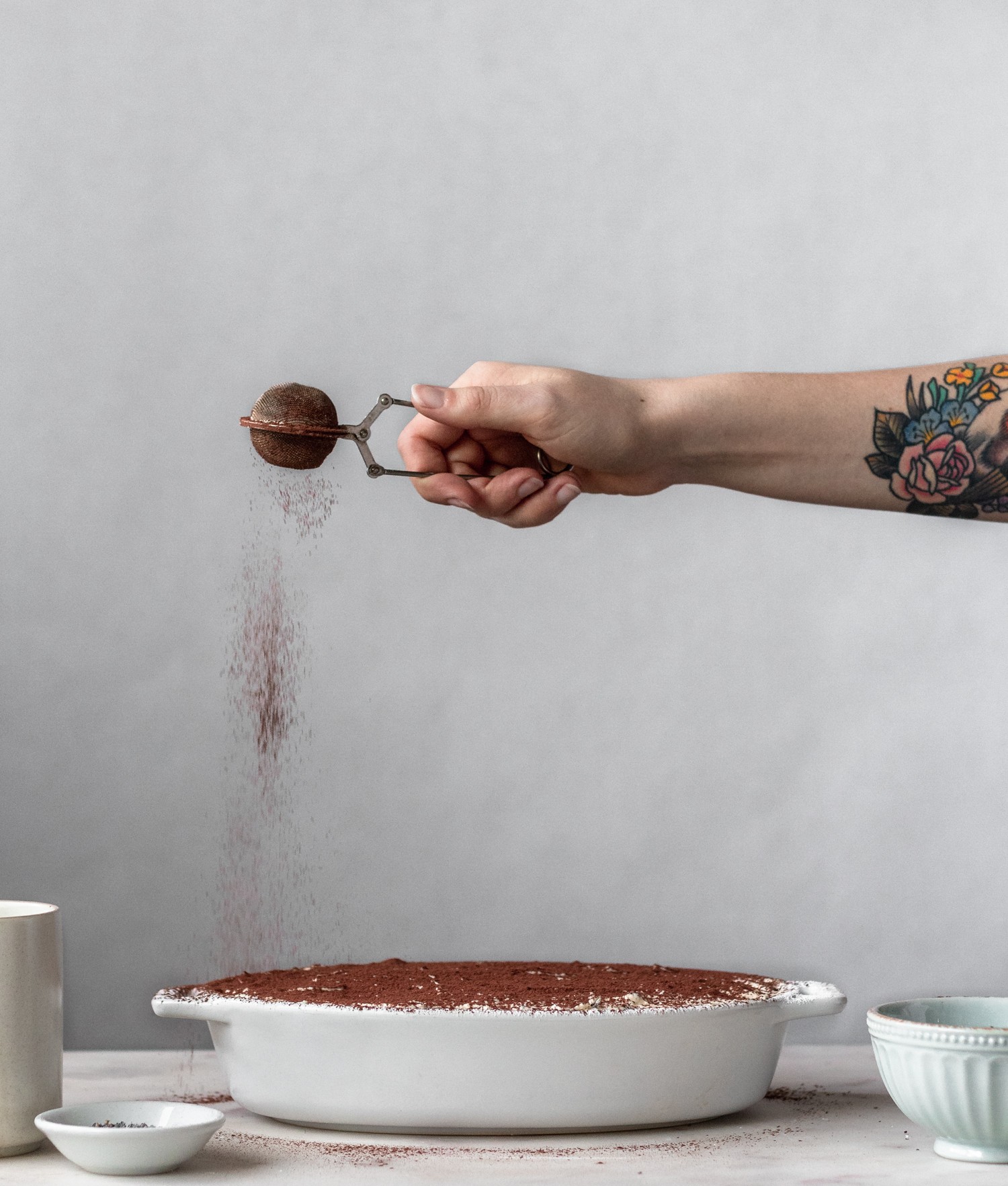 A woman's arm dusting cocoa powder over a white dish with London fog tiramisu with a white background.