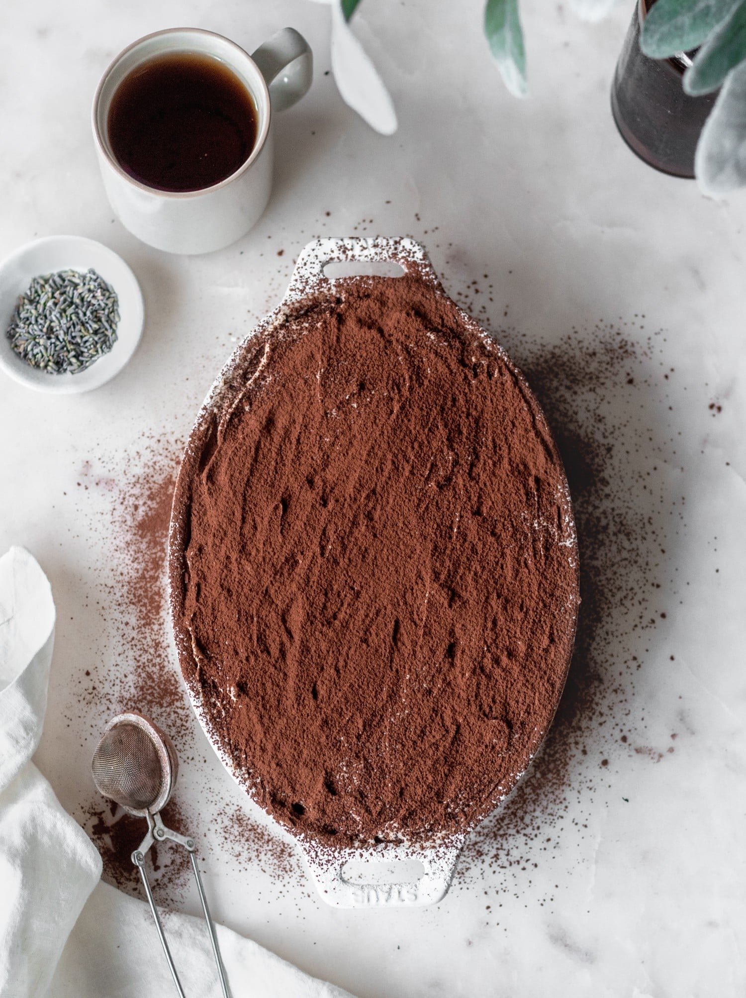 An overhead image of a white tray of custard dusted with cocoa powder on a white counter next to a small sifter, cup of tea, and white bowl of lavender.