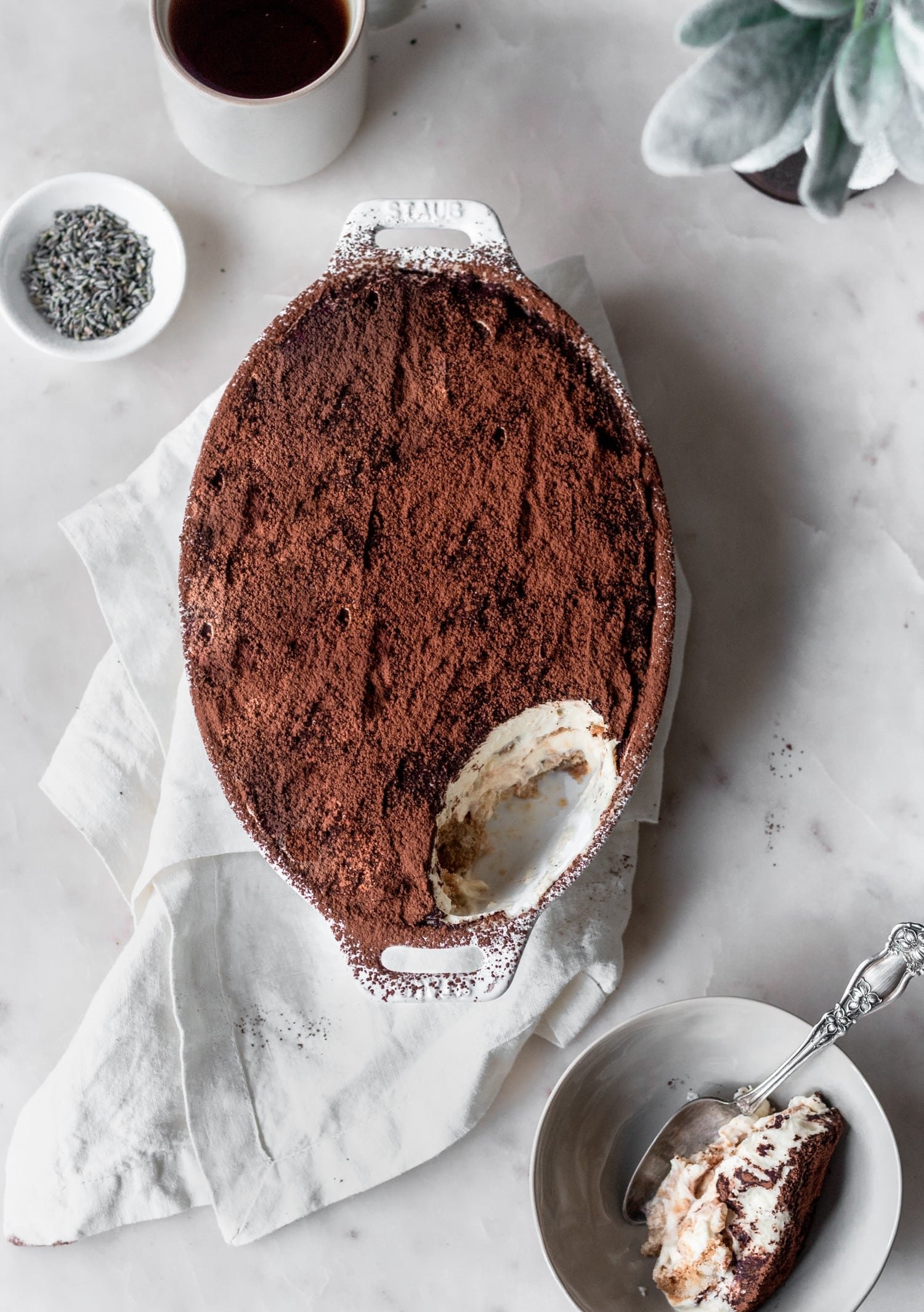 An overhead image of a white oval dish with London fog tiramisu with a scoop taken out of it on a marble counter next to a dish of tiramisu and two cups of tea.