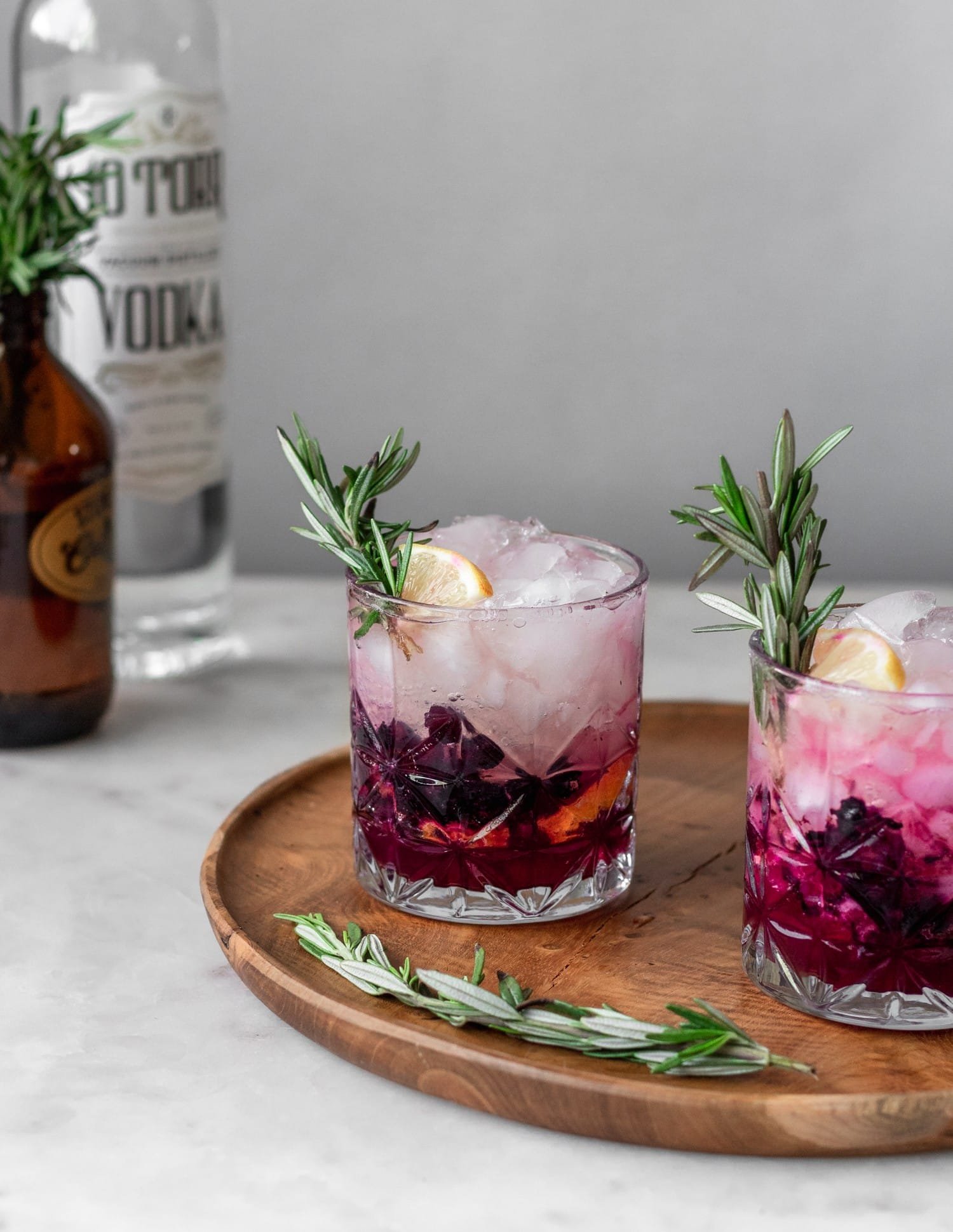 Two blueberry lemon vodka spritz cocktails on a wood board next to rosemary and a bottle of vodka on a marble background.