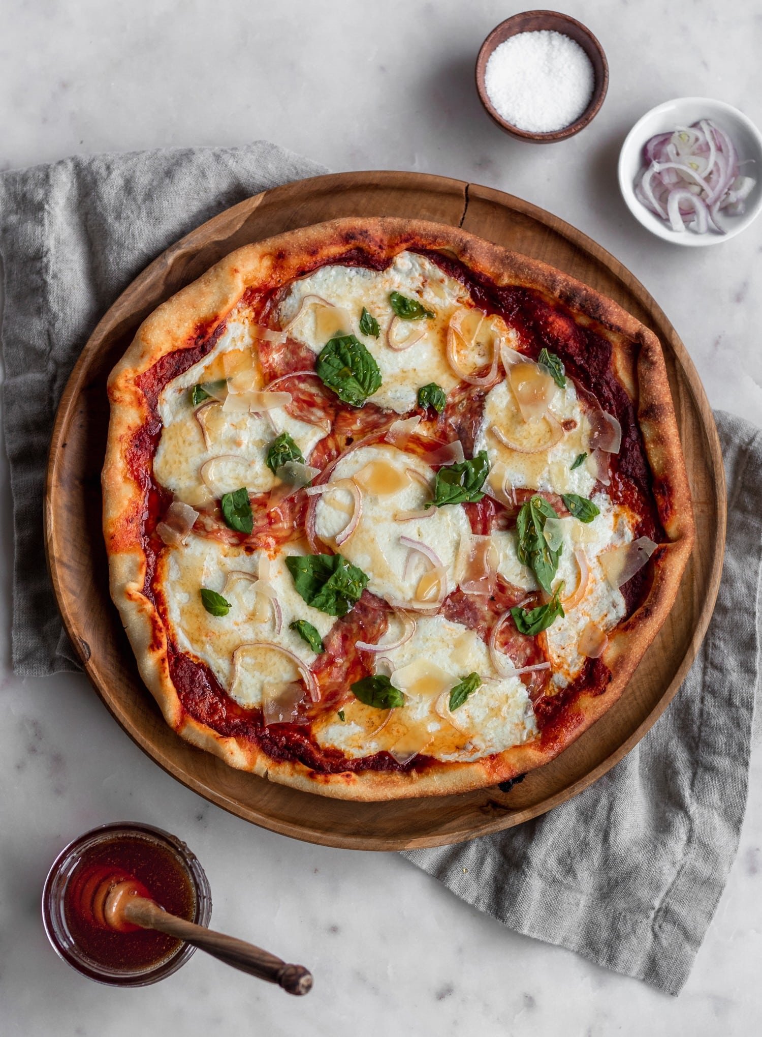 An overhead photo of pizza with soppressata on a wood tray next to a tan linen and jar of hot honey with a white marble background.