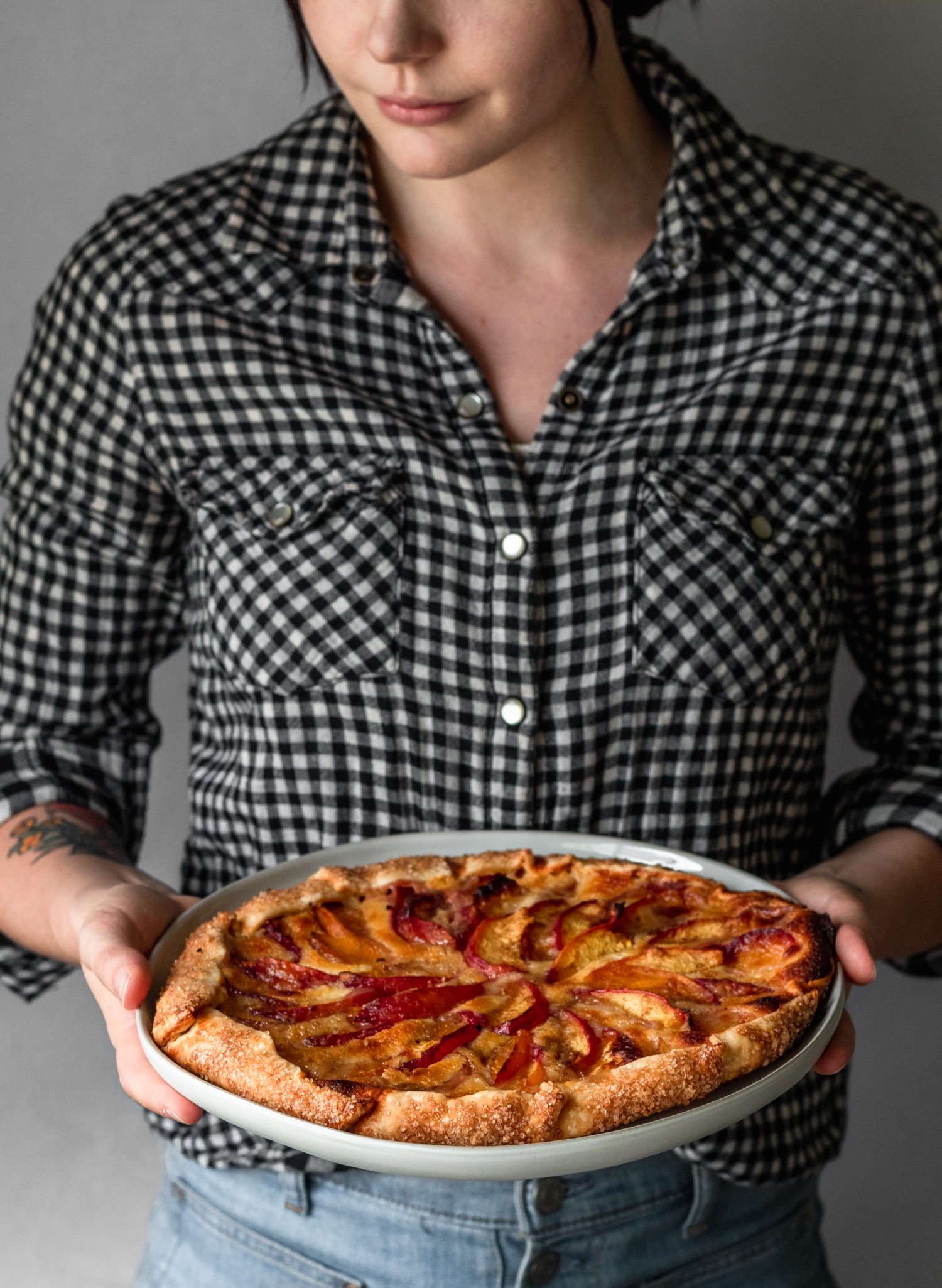 A side image of a woman wearing a black and white gingham shirt holding a nectarine galette on a white plate.