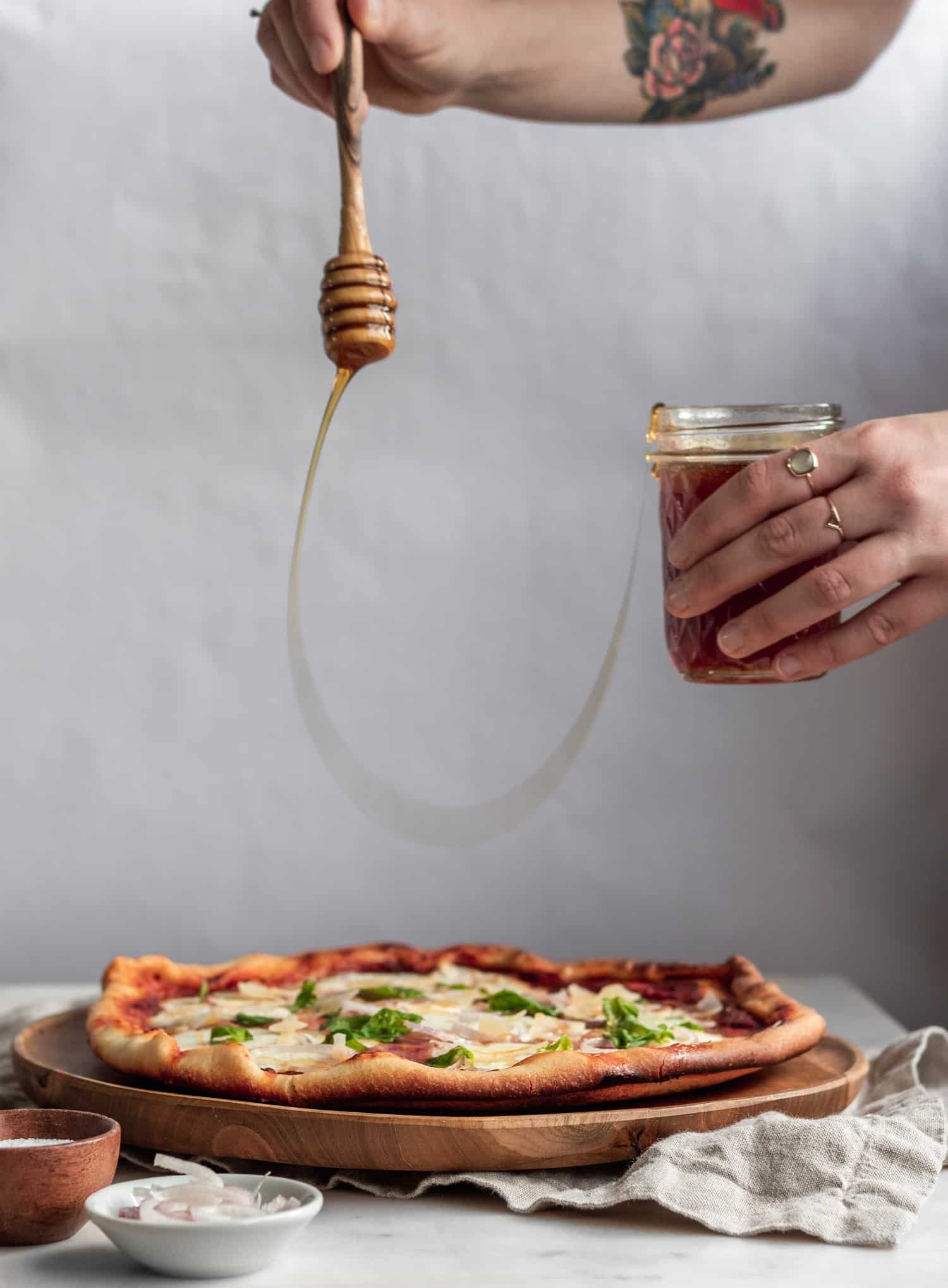 A woman's hand drizzling hot honey over a pizza with a white background.