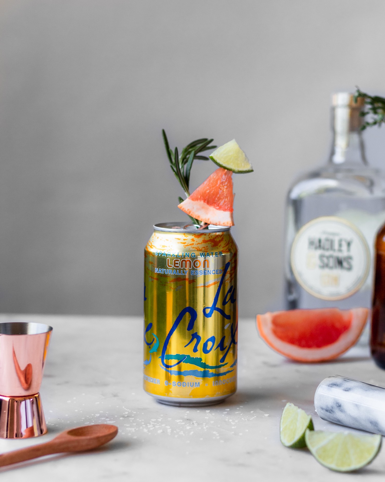 A side image of a lemon can of sparkling water with citrus garnish on a marble counter next to citrus wedges, gin, and a copper shot glass.