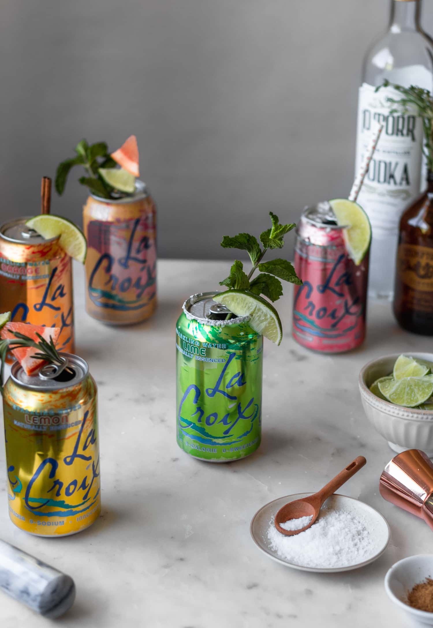 A side image of five cocktails in a can on a marble counter surrounded by cocktail garnishes, alcohol bottles, and a plate of salt.