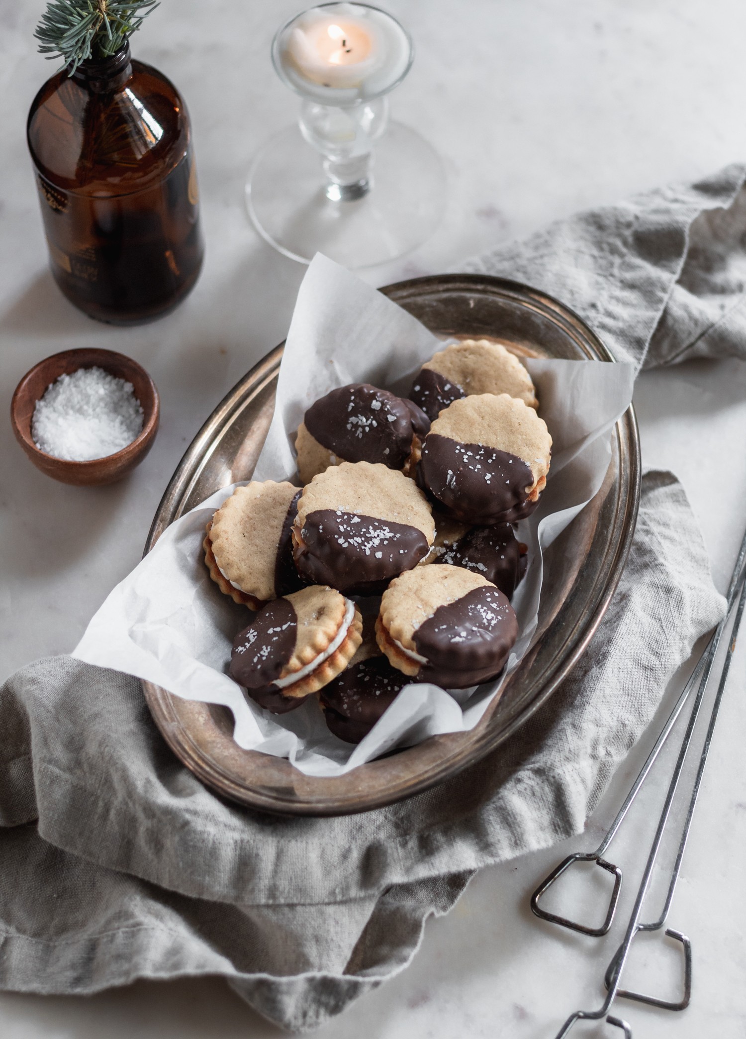 S'mores sandwich cookies in a metal dish on a grey linen next to skewers and a wood bowl of salt with a marble background.