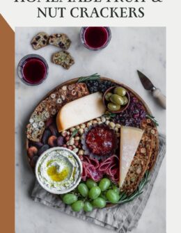 An overhead image of a cheese board with homemade fruit and nut crisps on a white marble counter next to a beige linen, glasses of red wine, and a cheese knife.