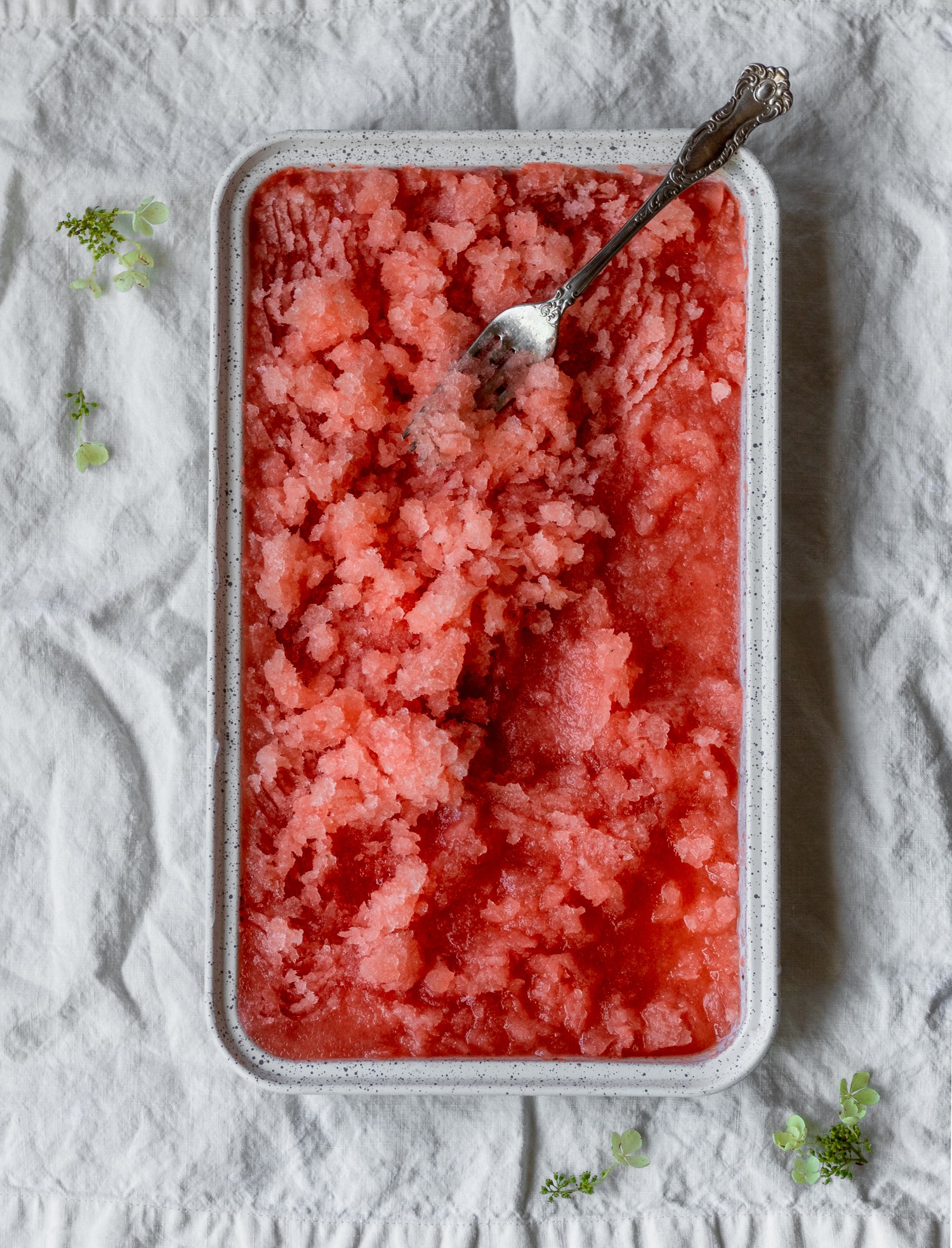 An overhead image of strawberry aperol spritz granita in a white tin on a white linen.