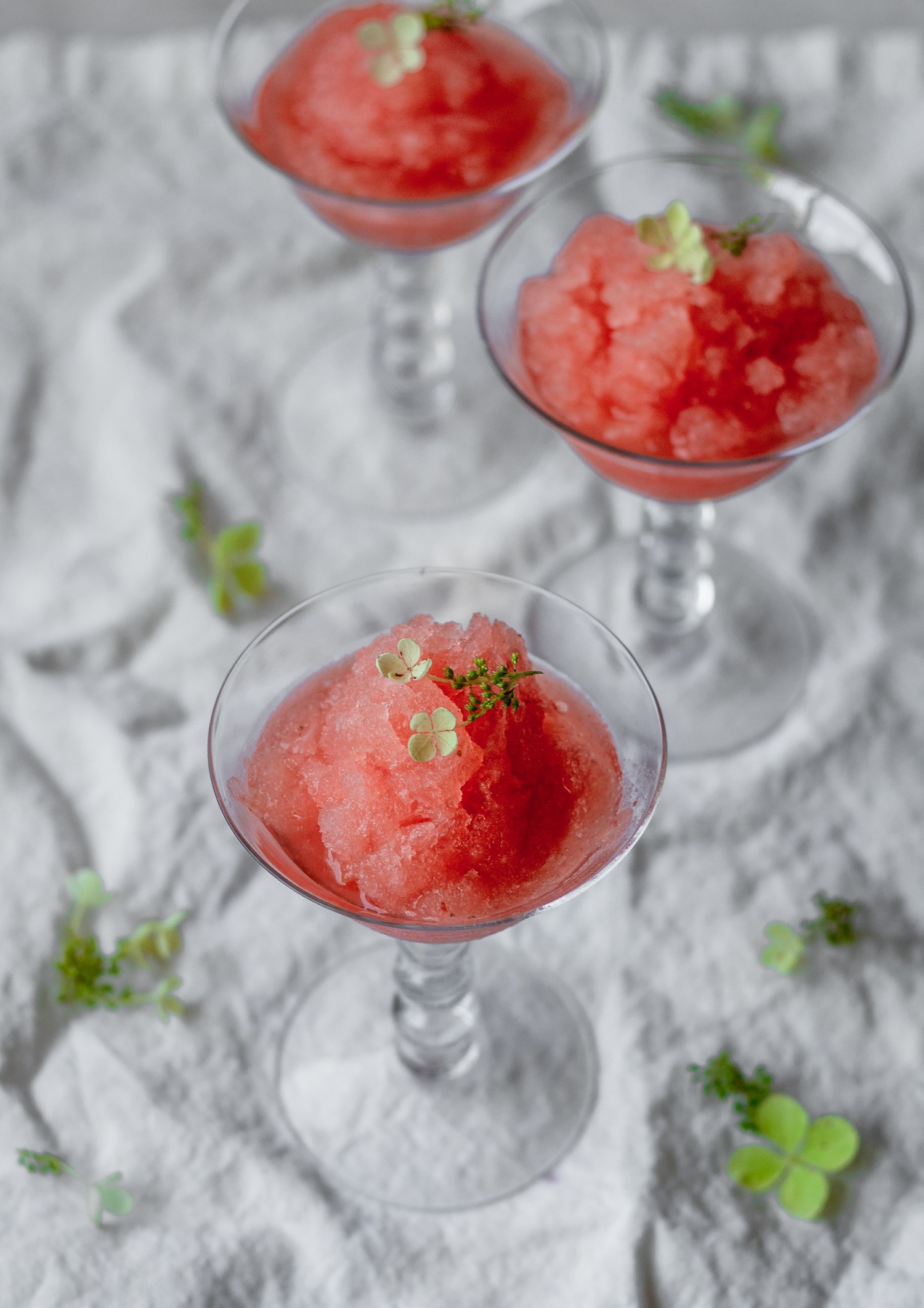 A side image of three glasses with strawberry aperol sprtiz granita on a white linen surrounded by green blossoms.