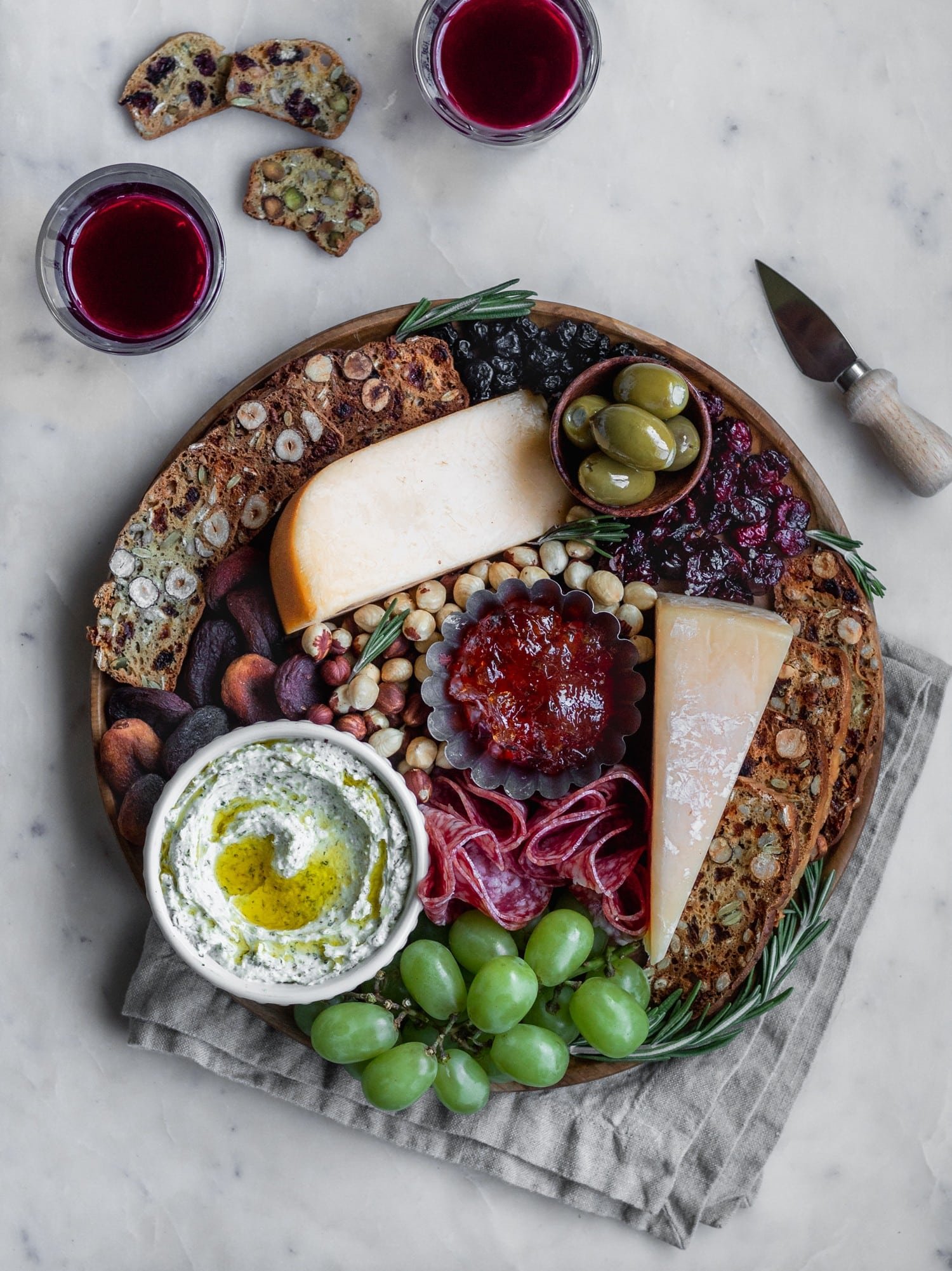 An overhead image of a cheese board with homemade fruit and nut crisps on a white marble counter next to a beige linen, glasses of red wine, and a cheese knife.