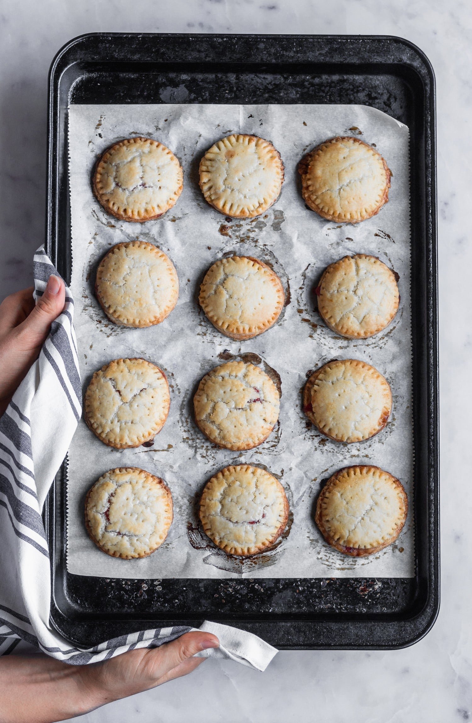 An overhead image of a woman holding a sheet pan with baked strawberry balsamic hand pies over a white marble counter.