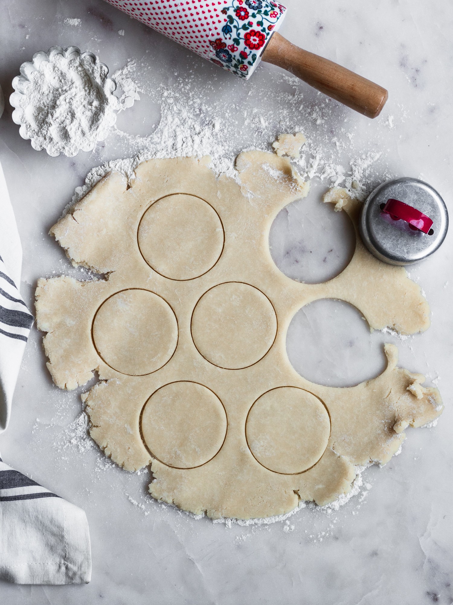 An overhead photo of pie dough with rounds cut out of it on a marble counter next to a bowl of flour and rolling pin.