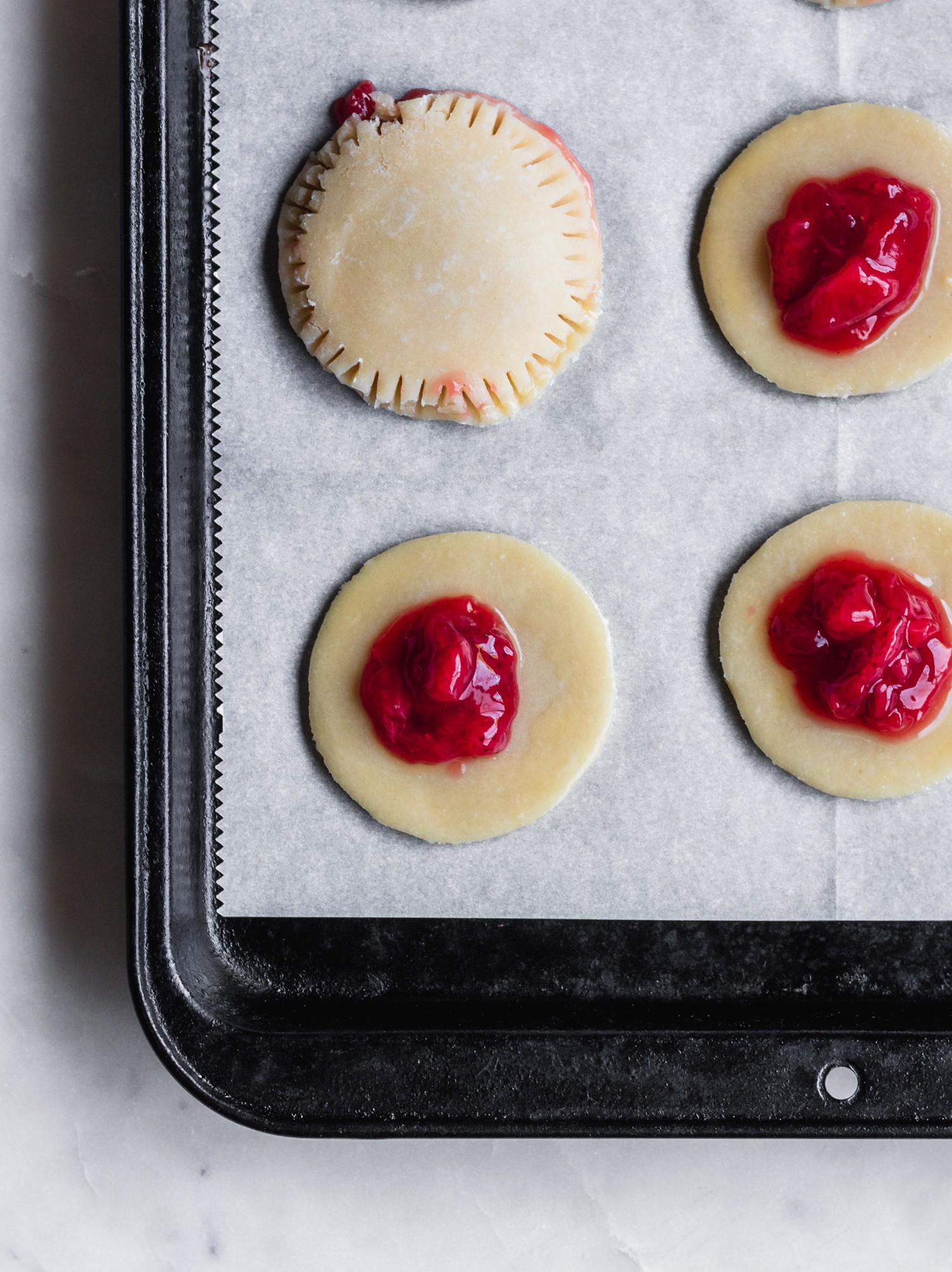 A closeup photo of pie dough rounds with strawberry jam in the middle on parchment paper.