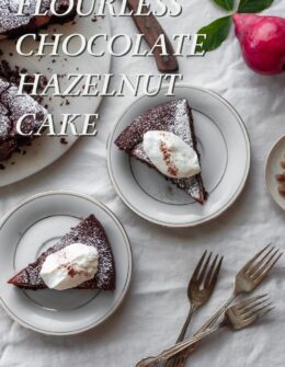 An overhead image of two white plates with chocolate cake slices and whipped cream on a white linen next to the whole cake, vintage forks, and a red pear.