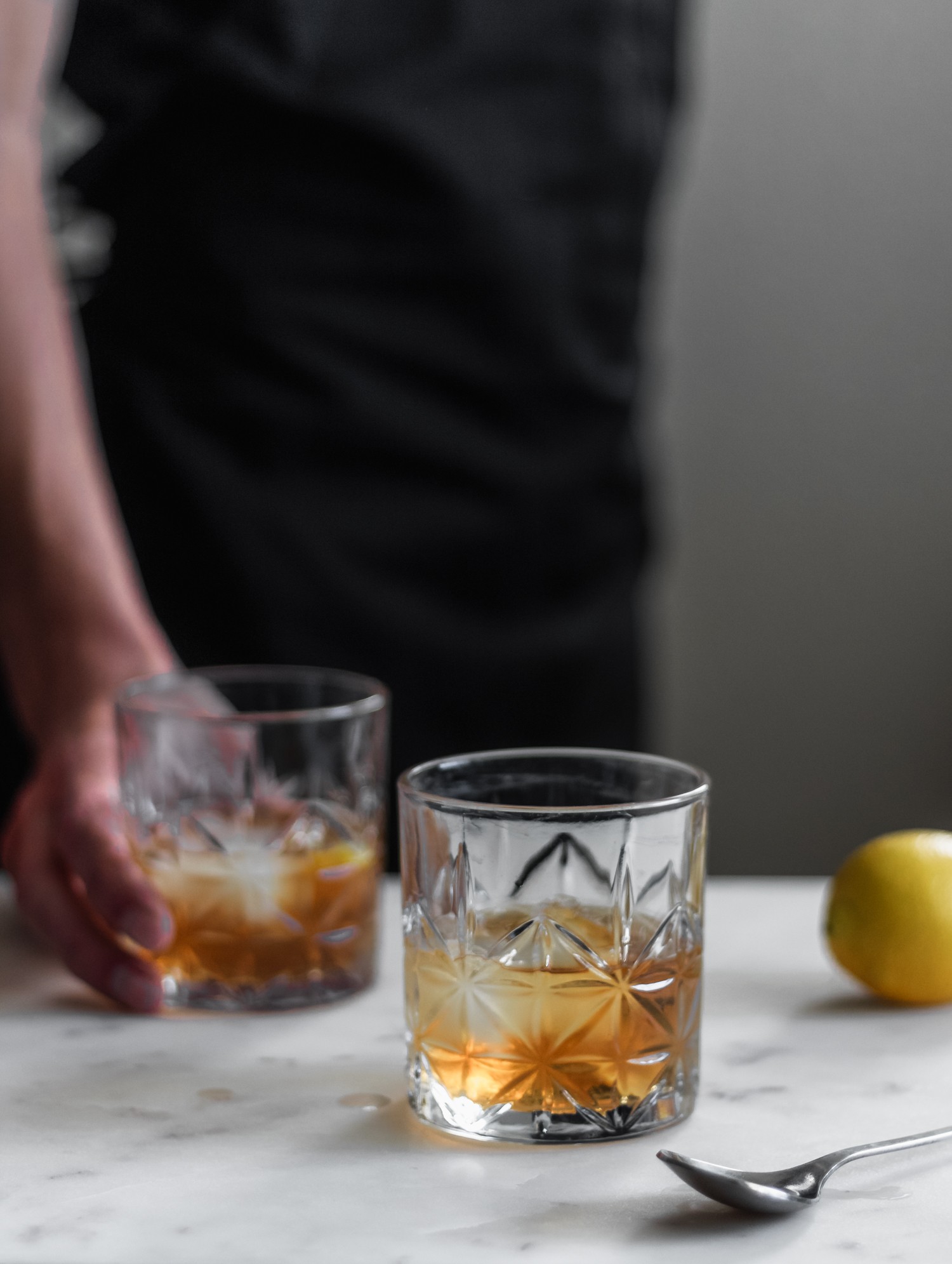 A closeup photo of a ginger old fashioned on a marble counter with a man in black holding another cocktail in the background.