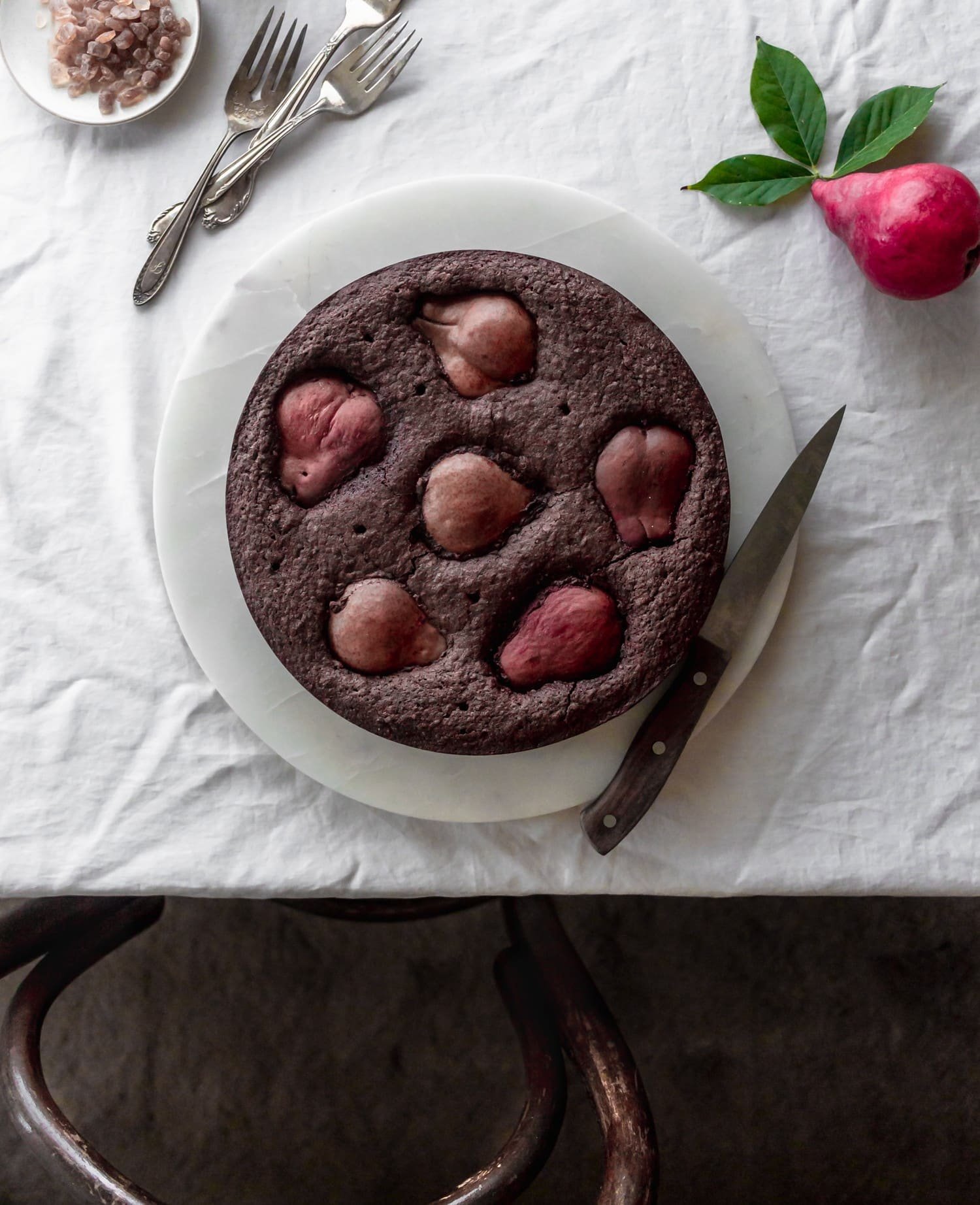 An overhead image of a chocolate cake with pears on a white marble cake plate on a white linen next to a wood knife and red pear.