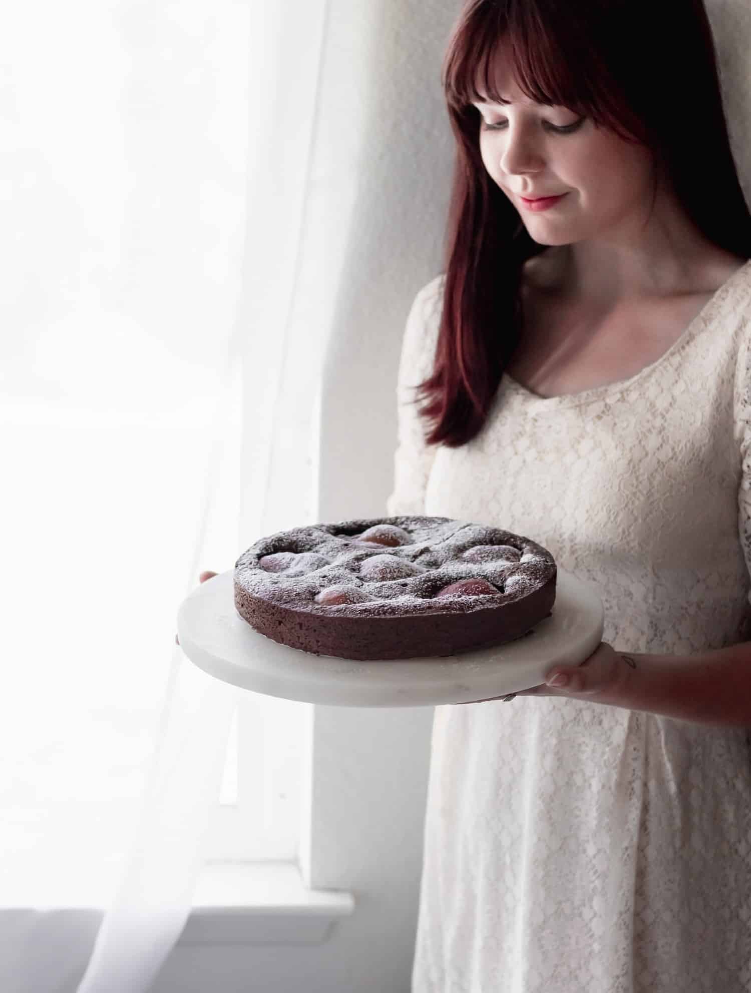 A side image of a woman wearing a white lace dress holding a Torta Caprese in front of a window with a white curtain.